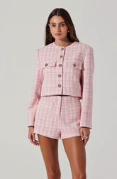 A woman is wearing a pink and white checkered tweed blazer with long sleeves and a round neckline, featuring decorative gold buttons down the front and on the chest pockets. She is also dressed in matching pink and white checkered shorts.